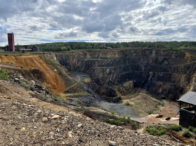 Großer Tagebau in einer Schlammebene umgeben von Bäumen, Gebäuden und einem Schuppen, mit bewölktem Himmel und Boden bedeckt mit Steinen und Pflanzen.