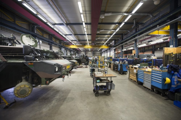 Großes Lagerhaus mit Maschinen, Fahrzeugen auf der linken Seite, Transportwagen mit Gegenständen und Holzboxen auf der rechten Seite und Deckenbeleuchtung.