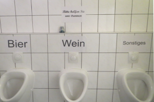 Drei weiße Toiletten an einer Wand montiert mit Schildern darüber, die jeweils 'Bier Wein' und 'Sonstiges' lesen.