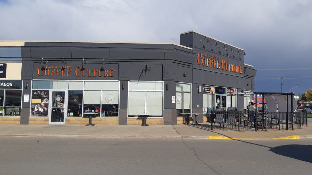 Ein Kaffeeladen an einer Straßenecke mit Fahrzeugen, Bäumen und einem Gebäude im Hintergrund unter einem blauen und weißen Himmel.