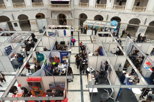 Große Gruppe von Menschen auf einer internationalen Fachmesse mit Ständen, Bannern, Tischen, Stühlen, Luftballons und einem Gebäude mit Bögen, Säulen, Geländern und einem Hintergrundbild.