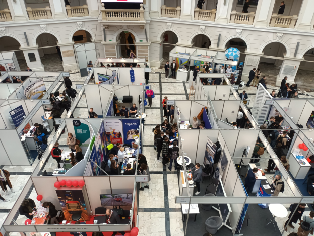 Große Gruppe von Menschen auf einer internationalen Fachmesse mit Ständen, Bannern, Tischen, Stühlen, Luftballons und einem Gebäude mit Bögen, Säulen, Geländern und einem Hintergrundbild.