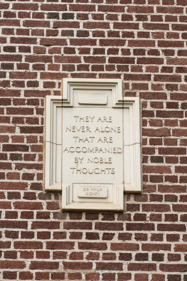 Eine Steinplatte, die auf einer Backsteinwand mit der eingravierten Inschrift "Sie sind nie allein, die von edlen Gedanken begleitet werden" in fetter Schrift montiert ist.