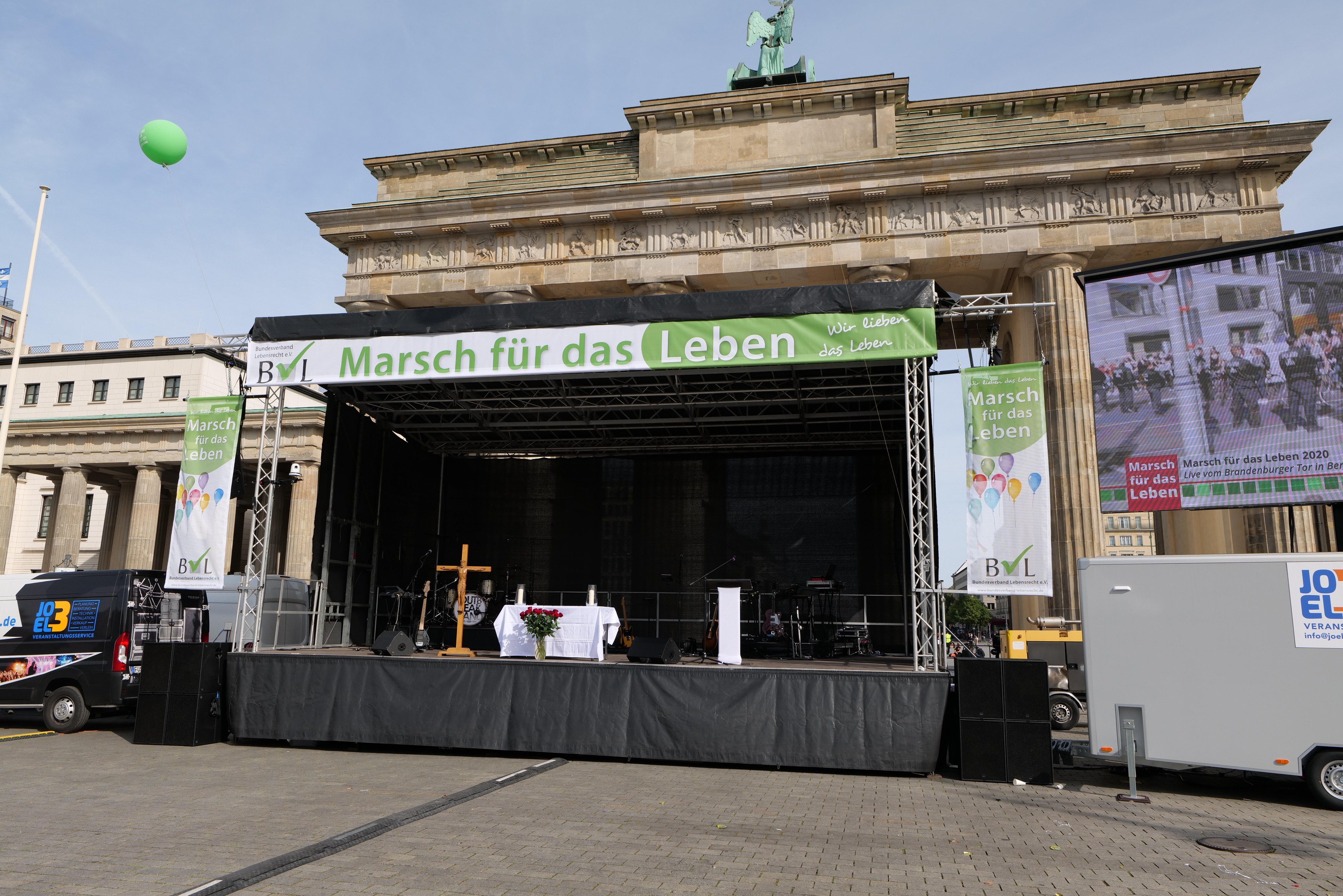 Bühne vor dem Brandenburger Tor mit einem Tisch, Bannern, Lautsprechern, Fahrzeugen, Gebäuden, einer Statue, einem Fahnenmast und Wolken am Himmel.