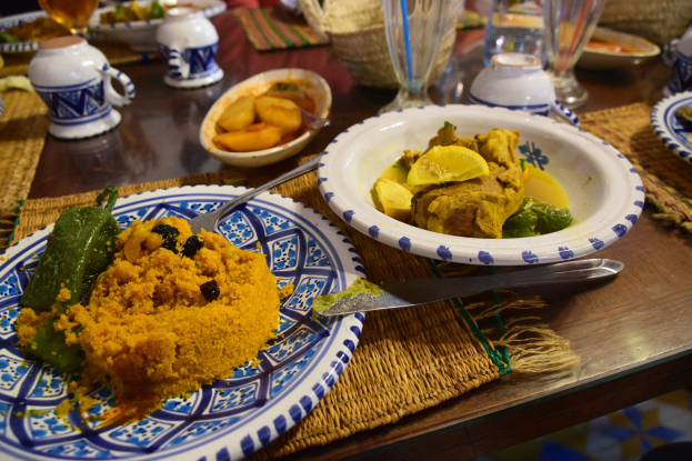Ein Tisch mit Tellern, Gläsern, Schüsseln, Krügen, Bestecken und Matten, mit Menschen im Hintergrund bei einem traditionellen marokkanischen Essen.