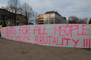 Eine Gruppe von Menschen hält ein Schild mit der Aufschrift 'Rechte für alle Menschen Stoppt Polizeigewalt' auf dem Boden stehend mit einem Straßenschild, einem Schild, Bäumen, Gebäuden mit Fenstern und einem bewölkten Himmel im Hintergrund.