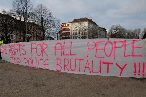 Eine Gruppe von Menschen hält ein Schild mit der Aufschrift 'Rechte für alle Menschen Stoppt Polizeigewalt' auf dem Boden stehend mit einem Straßenschild, einem Schild, Bäumen, Gebäuden mit Fenstern und einem bewölkten Himmel im Hintergrund.