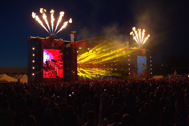 Eine große Menschenmenge auf einem Musikfestival mit einer hell erleuchteten Bühne mit Künstlern und Bannern, Zelte links und Feuerwerk am dunklen Himmel.