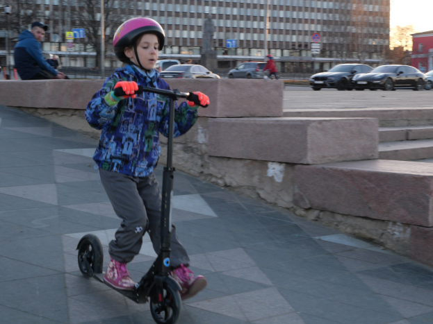 Ein junger Junge mit Helm und Handschuhen fährt einen Roller eine Treppe hinunter, mit Fahrzeugen, Menschen, Bäumen, Pfosten, Brettern, Gebäuden und einem klaren blauen Himmel im Hintergrund.