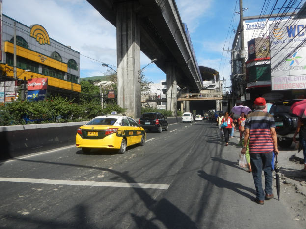 Ein gelbes Taxi fährt auf einer Stadtstraße mit hohen Gebäuden, Passanten mit Regenschirmen auf dem Gehweg, einer Brücke im Hintergrund und einem bewölkten Himmel mit Strommasten und Drähten.