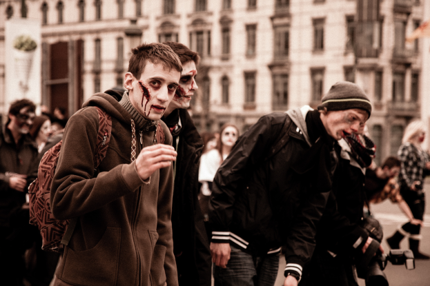 Eine Gruppe von Menschen in Zombie-Kostümen geht eine Straße entlang, einige tragen Kostüme und einer hält eine Kamera, und ein Gebäude im Hintergrund.