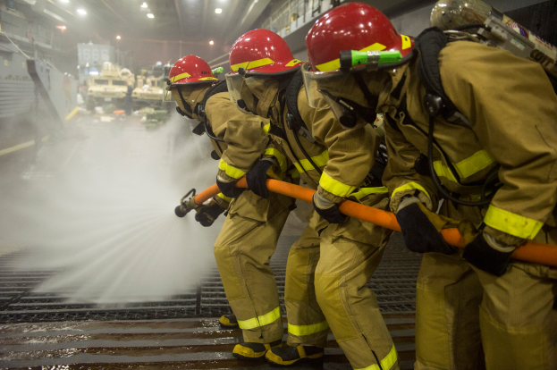 Feuerwehrleute in Helmen und Handschuhen halten Rohre und sprühen Wasser auf ein Feuerwehrauto, mit Lichtern, Geländern und Tafeln mit Text im Hintergrund.