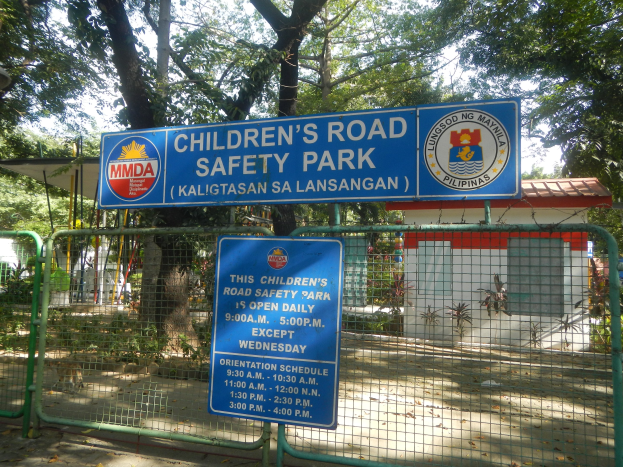 Metallzaun um einen Kinderradweg-Sicherheitspark mit zwei blauen Informationsschildern, einem kleinen Haus mit Dach, Pflanzen, Bümern und einem bewülkerten Himmel.