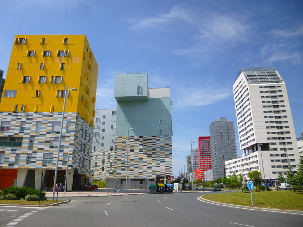 Eine Stadtstraße mit hohen Gebäuden im Hintergrund, Straßenlaternen, Autos, Informationsschildern, Büschen, Bäumen und einem bewölkten Himmel, mit Gebäuden der Universität Rotterdam mit einzigartiger Architektur.
