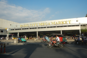 Ein belebter Pasig City Mega Markt mit Fahrzeugen auf der Straße, Menschen, die herumlaufen, einem Gebäude mit Text im Hintergrund und einer bewölkten Himmel.