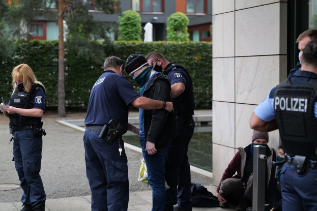 Eine Gruppe von Polizisten vor einem Gebäude mit einer Frau, die ein Buch und einen Stift hält, während Menschen in der Nähe sitzen und stehen, mit Bäumen und Gebäuden im Hintergrund.