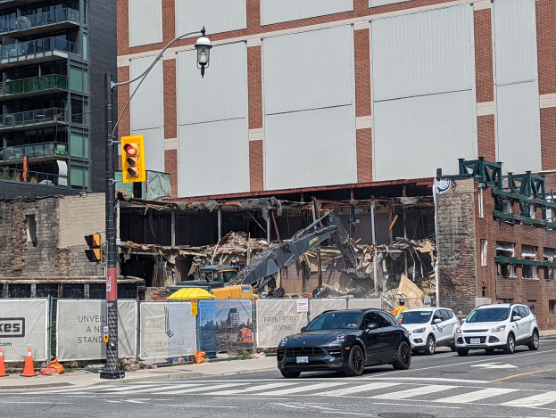 Ein großes Gebäude wird in der Mitte einer Straße abgerissen, mit Fahrzeugen, Verkehrskegeln, einer Ampel und einem Kran im Vordergrund und Gebäuden mit Fenstern im Hintergrund unter einem sichtbaren Himmel.