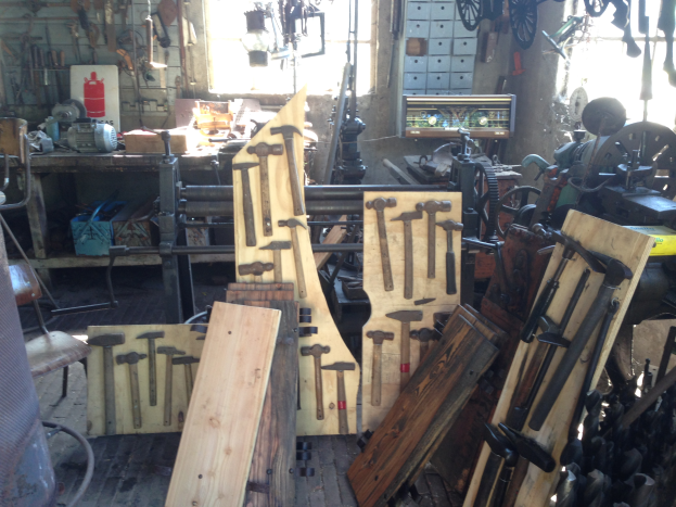 Ein mit verschiedenen Werkzeugen, Maschinen und Holzplanken gefüllter Werkstattraum, mit einem Stuhl links, Werkzeugen an der Wand und einem Fenster im Hintergrund.