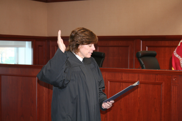 Eine Frau in einer schwarzen Robe steht vor einem Richtertisch und hält eine Akte in der Hand, mit einem Mikrofon und einer Flagge auf dem Tisch und Stühlen und einem Fenster dahinter.