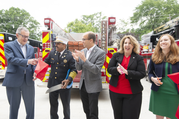 Gruppe von Menschen bei einer Einweihungszeremonie für die Alexandria Fire Department, die vor einem Feuerwehrauto klatschen und lächeln, mit Bäumen und einem klaren blauen Himmel im Hintergrund.