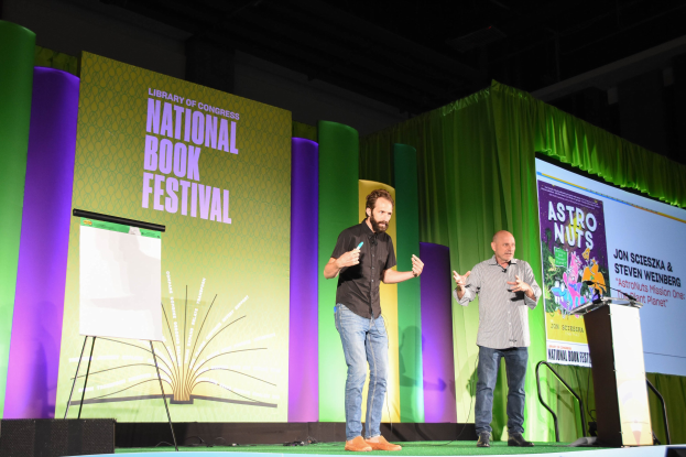 Zwei Männer auf einer Bühne beim Nationalen Buchfestival, mit einem Podium und einem Mikrofon rechts daneben und einem großen Bildschirm dahinter.