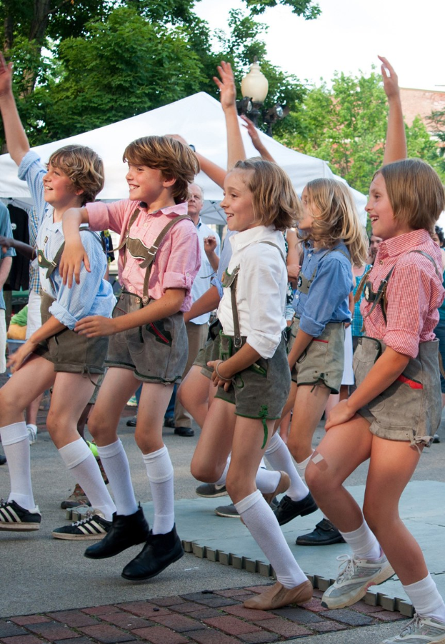 Kinder in traditioneller bayrischer Tracht, die auf einer Straße tanzen, umgeben von einer Menge, Zelten und Gebäuden unter einem klaren blauen Himmel.
