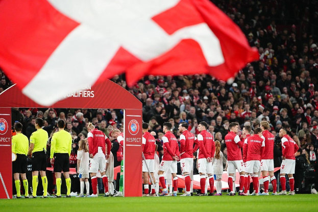 Gruppe von Menschen auf einem Fussballfeld mit einer roten und weißen Flagge im Vordergrund, einem Bogen mit der Aufschrift "Bayern Munich vs Bayern Munich Wetten & Vorschau" im Hintergrund und einer großen Menge im Stadion.