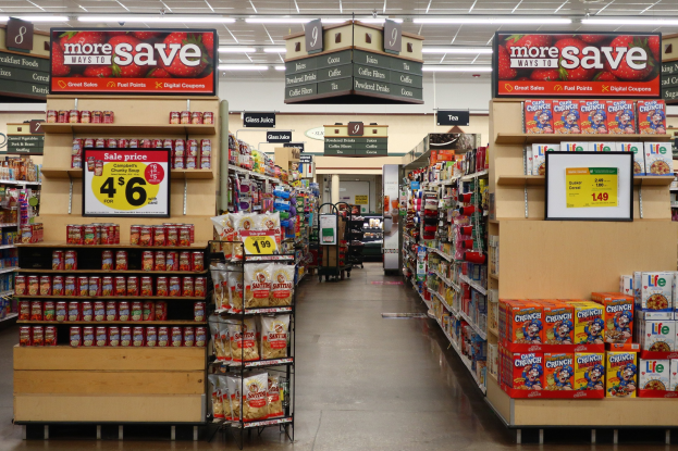 Innensicht eines Lebensmittelgeschäfts mit verschiedenen Lebensmitteln auf Regalen und dem Boden, beleuchtet von Deckenlampen, mit Informationsschildern und einer Person im Hintergrund.