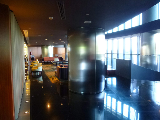 Eine geräumige First-Class-Lounge am Hong Kong International Airport mit großen Fenstern, hohen Decken mit elektrischen Leuchten, Sitzgelegenheiten wie Stühle und Sofas, Tischen, einem Teppichboden und einer zentralen Säule.