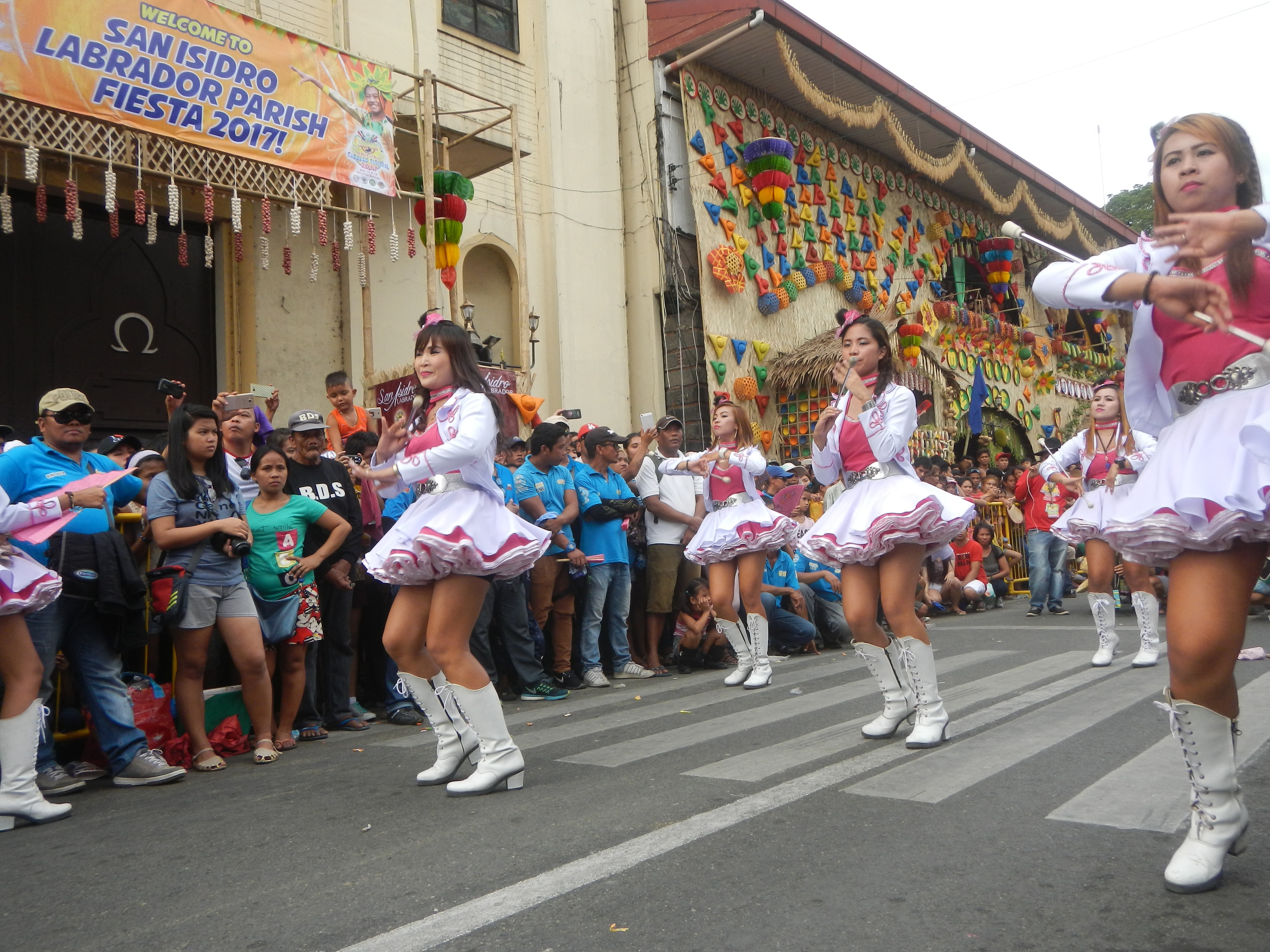 Eine Gruppe von Frauen in pinken und weißen Outfits tanzt auf der Straße, umgeben von einer Menge Menschen, die stehen und zuschauen, mit Gebäuden, Bäumen und einem klaren blauen Himmel im Hintergrund.