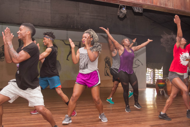 Gruppe von Menschen, die in einem Studio mit einer bemalten Wand, einem Fenster und Deckenbeleuchtung tanzen, wahrscheinlich während eines Zumba-Kurses.