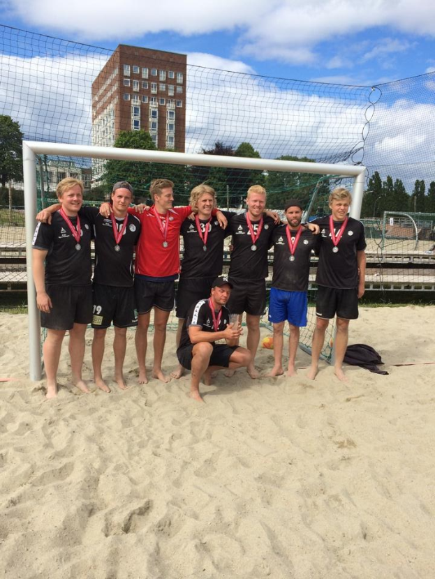 Mannergruppe in Sportbekleidung auf einem sandigen Strand, die Medaillen tragen und von einem Netz umgeben sind, mit Bäumen, Gebäuden und einem bewölkten Himmel im Hintergrund.