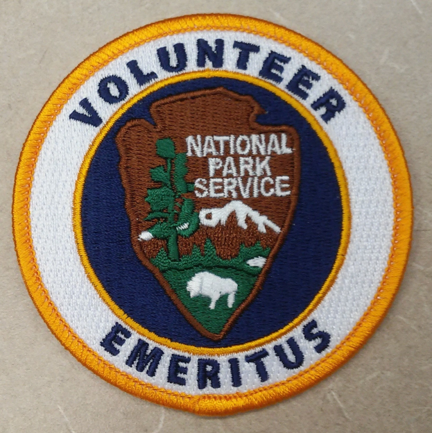 Runder Flicken mit blauem Rand, weißem Hintergrund und gelbem inneren Kreis, der in der Mitte die fett gedruckten schwarzen Buchstaben "Volunteer National Park Service Emeritus" zeigt.