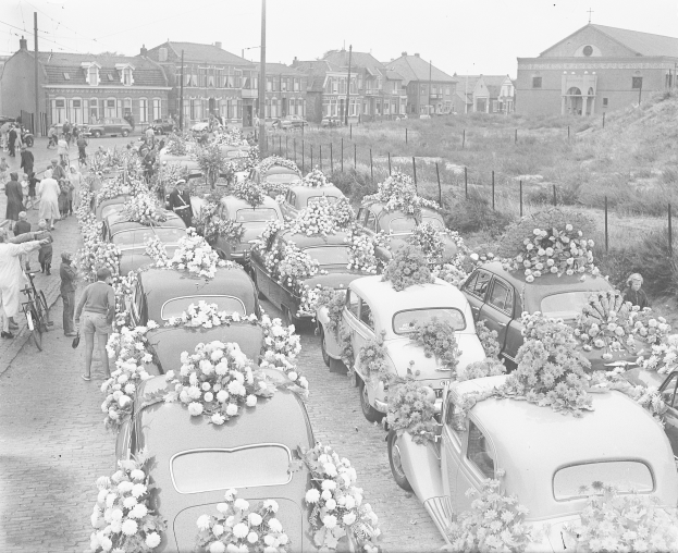 Schwarzes und weißes Bild einer Beerdigungsprozession mit mehreren Autos, die mit Blumenbouquets geschmückt sind, umgeben von Menschen, einige halten Fahrräder, mit Gebäuden, Pfählen und einem klaren Himmel im Hintergrund.