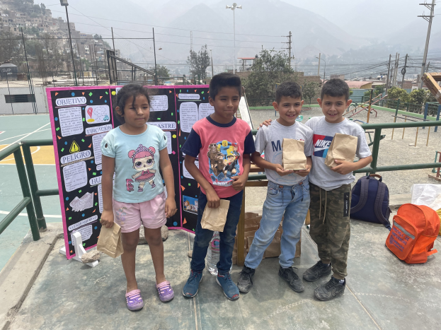 Eine Gruppe von Kindern steht auf einem Basketballfeld und hält Papierbeutel in den Händen, mit Beuteln und Gegenständen rechts und einer Tafel mit Text links, vor einem Hintergrund aus Zaun, Pfosten, Straßenlaternen, Bäumen, Gebäuden, Hügeln und Himmel.