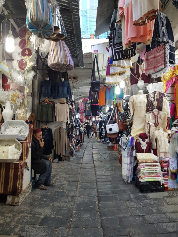 Ein belebter Straßenmarkt in der Altstadt von Jerusalem mit Ständen, die Kleidung, Taschen, Lampen und andere Artikel verkaufen, während Menschen im Hintergrund durch die Stände schlendern und einkaufen.