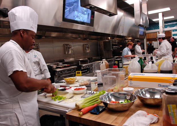 Eine Gruppe von Köchen in einer Küche bei der Zubereitung von Speisen auf einem Schneidbrett, wobei ein Koch im Vordergrund in einer weißen Schürze und Mütze steht und ein Messer hält und von Utensilien, Schüsseln, Gemüse, einem Handy, Flaschen und anderen Gegenständen umgeben ist, mit einem Fernseher und Deckenleuchten im Hintergrund.