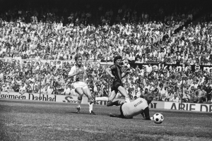 Schwarzes und weißes Foto von Männern, die auf einem Feld Fußball spielen, mit Zuschauern im Hintergrund und einer Bundesliga 1978-79 Ajax 1-1 Hannover 96 Fahne.