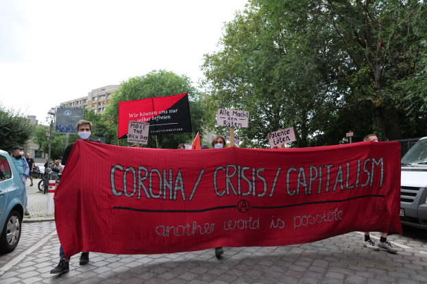 Eine Gruppe von Menschen marschiert mit einer roten Fahne, auf der 'Corona-Krise/Kapitalismus' steht, eine Straße entlang. Im Hintergrund sind geparkte Fahrzeuge, Gebäude und Bäume unter einem klaren blauen Himmel zu sehen.