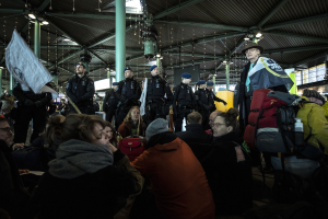 Eine Gruppe von Menschen vor einer Menge an einem Bahnhof stehend, einige tragen Mützen und tragen Taschen, im Hintergrund Säulen und Deckenleuchten; die Menge scheint zu protestieren, hält Schilder und Banner.