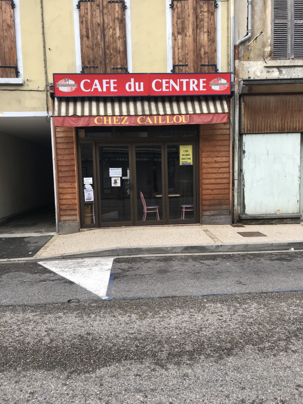 Außenansicht eines Gebäudes mit einer 'Cafe du Centre'-Tafel, Fenstern, einer Tür und einer Straße davor mit einer Texttafel.