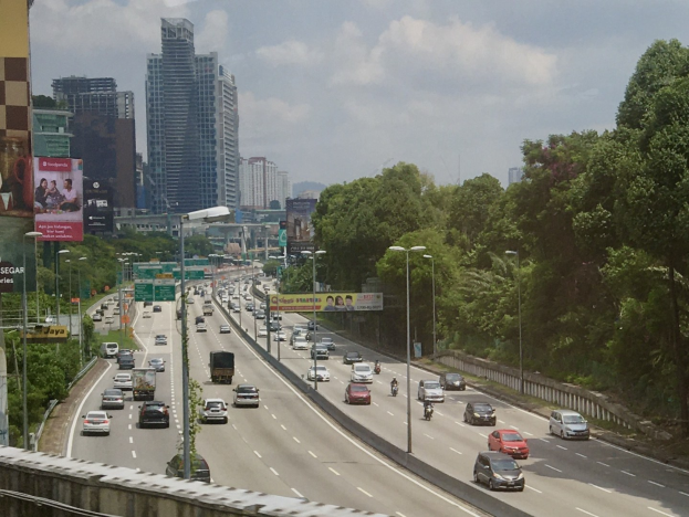 Starker Verkehr auf einer Autobahn neben hohen Gebäuden, Straßeninfrastruktur und Bäumen unter einem bewölkten Himmel.