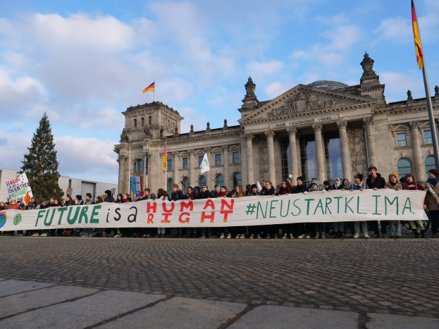 Eine Gruppe von Menschen steht vor dem Reichstaggebäude in Berlin und hält ein Banner mit der Aufschrift "Zukunft ist ein Menschenrecht". Das Gebäude hat Säulen, Fenster und Bögen und ist von Bäumen und Fahnenmasten umgeben. Der Himmel ist bewölkt.