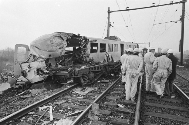 Ein Schwarz-Weiß-Bild von einer Zugkollision auf den Schienen mit Menschen in Helmen in der Nähe, umgeben von Strommasten, Bäumen, Gebäuden und einem klaren Himmel.