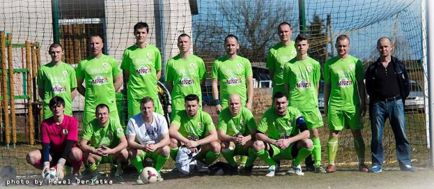 Eine Gruppe von Männern in grünen T-Shirts steht auf einem Fußballfeld, einige hocken, mit einem Ball vor ihnen in der Nähe eines Torpfostens; der Hintergrund umfasst Bäume, Gebäude und Fahrzeuge unter dem Himmel.