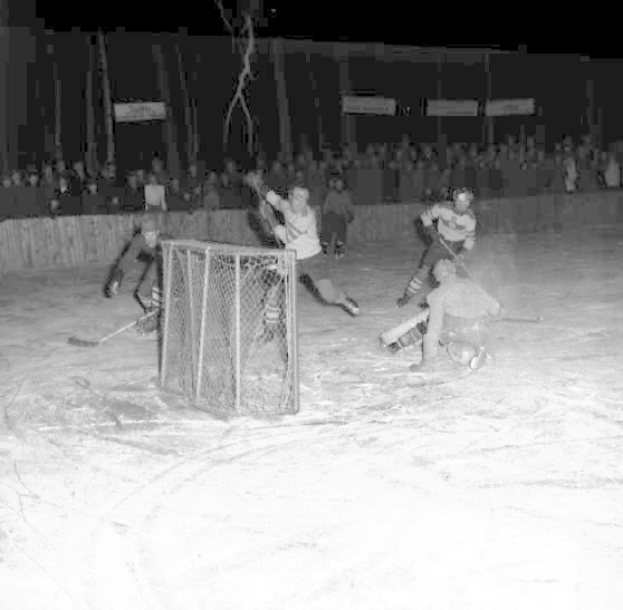 Schwarzes und weißes Foto eines Hockey-Spiels auf einem Eisstadion, mit einem Netz im Vordergrund und Zuschauern im Hintergrund.