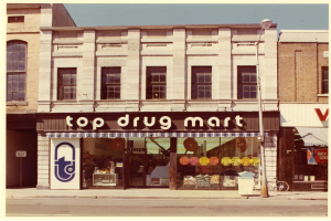 Ein Drogengeschäft an einer Stra√enecke mit einem Gebäude, einer Stra√enlaterne, einem geparkten Fahrrad, einem Müllcontainer und einem bewölktem Himmel.