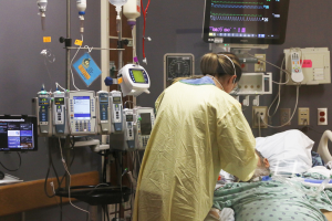 Ein Mann liegt in einem Krankenhausbett neben einem Monitor, mit einer Frau neben ihm; ein Tisch mit einem Monitor ist links und medizinische Geräte sind im Hintergrund zu sehen.