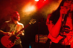 Eine Frau singt in ein Mikrofon auf einer hell erleuchteten Bühne, während ein Mann im Hintergrund eine Akustikgitarre spielt.