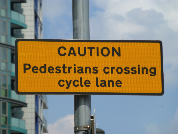 Gelbes Warnschild an einem Pfahl vor einem Gebäude, auf dem steht: "Fußgänger überqueren Fahrradspur".